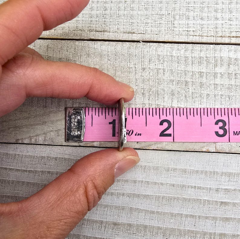 Puede incluir: Un anillo de plata se mide con una cinta m&eacute;trica rosa. El anillo se sostiene entre el pulgar y el &iacute;ndice, y la cinta muestra una medida de aproximadamente 2,5 cm. La cinta est&aacute; marcada con n&uacute;meros y l&iacute;neas negras.
