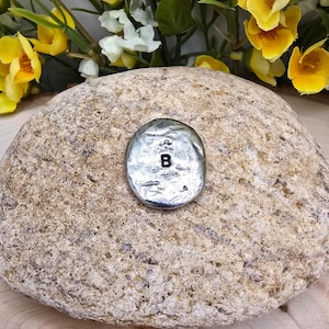 Puede incluir: Un peque&ntilde;o dije ovalado plateado con la letra "B" en negro. El dije est&aacute; colocado sobre una piedra grande, texturizada, de color marr&oacute;n claro. Flores amarillas y blancas en el fondo.