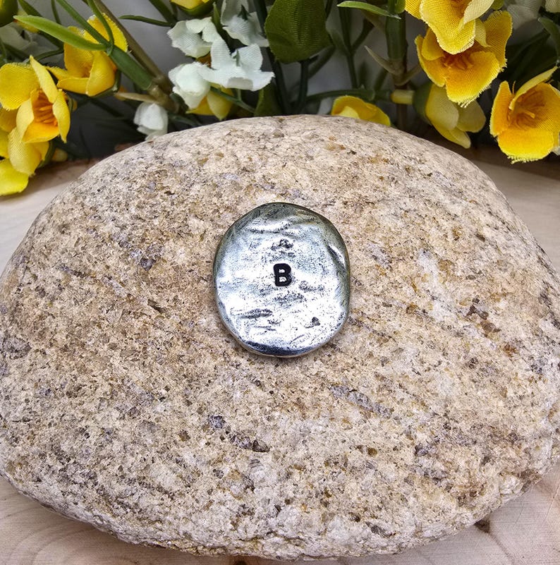 Puede incluir: Un peque&ntilde;o dije ovalado plateado con la letra "B" en negro. El dije est&aacute; colocado sobre una piedra grande, texturizada, de color marr&oacute;n claro. Flores amarillas y blancas en el fondo.