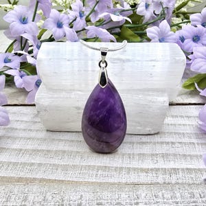 May include: A teardrop-shaped amethyst pendant on a silver chain. The pendant is a deep purple color and has a smooth, polished finish.