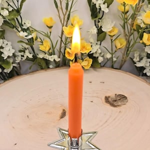 May include: A bright orange taper candle with a lit flame, held upright in a silver-toned metal star-shaped candle holder. The candle is positioned in the centre, featuring a smooth wax texture and a vibrant, solid orange colour throughout.