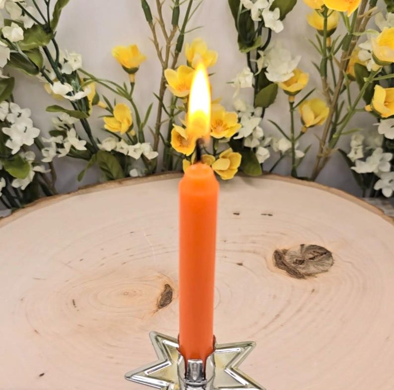 May include: A bright orange taper candle with a lit flame, held upright in a silver-toned metal star-shaped candle holder. The candle is positioned in the centre, featuring a smooth wax texture and a vibrant, solid orange colour throughout.