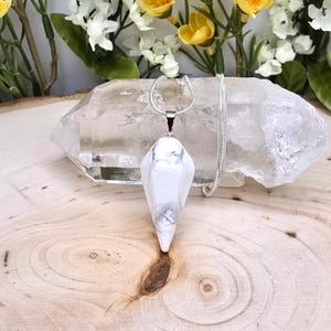 May include: A white and gray marbled stone pendant necklace with a silver chain. The pendant is a geometric shape, and the necklace is displayed on a clear crystal and a wooden surface.