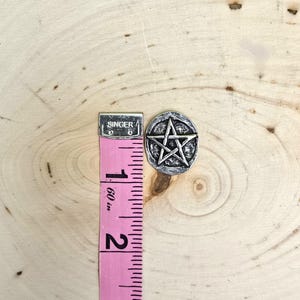Puede incluir: Un dije de pentagrama plateado, de aproximadamente 2,5 cm de di&aacute;metro, se muestra junto a una cinta m&eacute;trica rosa. El dije presenta una estrella de cinco puntas dentro de un marco circular. La cinta m&eacute;trica est&aacute; marcada en pulgadas.