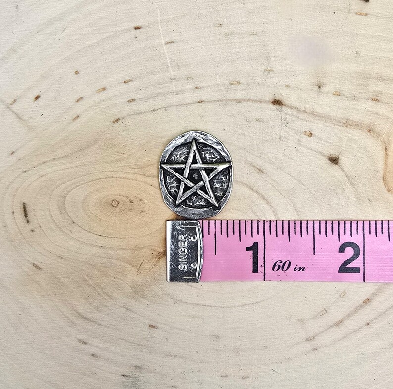 Puede incluir: Un colgante plateado de forma ovalada con un dise&ntilde;o de pentagrama en relieve. El colgante est&aacute; junto a una cinta m&eacute;trica rosa, que muestra una medida de aproximadamente 2,5 cm. El colgante tiene una superficie texturizada.