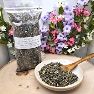 May include: A clear plastic bag filled with dried green nettle leaves, labeled Metaphysical Gypsy 1 oz Nettle Leaf, Cut Urtica Dioica, alongside a small white ceramic bowl containing a portion of the herbs and a wooden spoon.