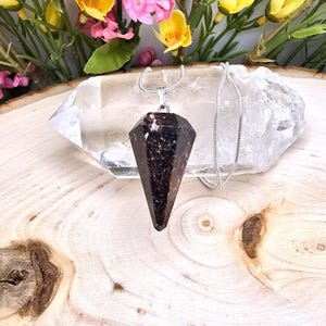 May include: A dark brown crystal pendulum pendant with a silver chain. The pendant is a faceted, elongated shape. The necklace rests on a clear quartz crystal and a wooden surface, with colorful flowers in the background.