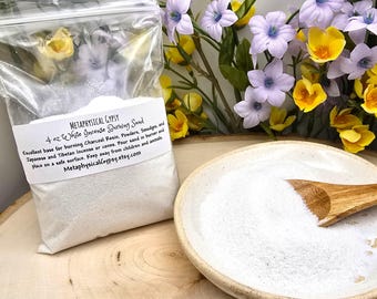 May include: A clear plastic bag of white incense burning sand with a label that reads "Metaphysical Gypsy". A small ceramic dish with sand and a wooden scoop is next to the bag. The background has yellow and purple flowers.