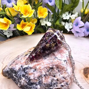 May include: A conical resin pendant featuring embedded dark purple garnet-like stones and gold-colored metallic flakes. The pendant is attached to a silver-toned metal chain and rests on a rough, multi-colored granite rock with pink, white, and black mineral flecks.