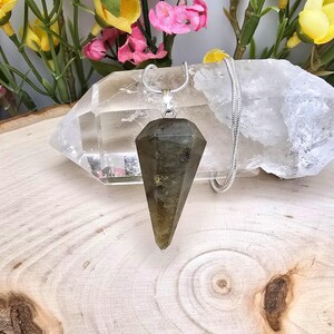 May include: A faceted, olive-green labradorite pendulum pendant hangs from a silver chain. The pendant is a long, pointed shape. The background includes a clear quartz crystal and colorful flowers on a wooden surface.