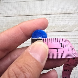 May include: A small, circular, vibrant blue object is positioned next to a pink measuring tape. The tape shows the object has a diameter of approximately 1.3 cm. The blue item has a smooth, solid texture and a rounded, flat-topped shape.