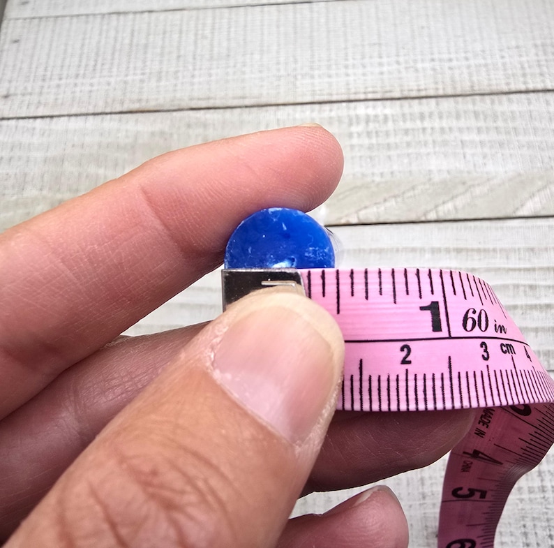 May include: A small, circular, vibrant blue object is positioned next to a pink measuring tape. The tape shows the object has a diameter of approximately 1.3 cm. The blue item has a smooth, solid texture and a rounded, flat-topped shape.