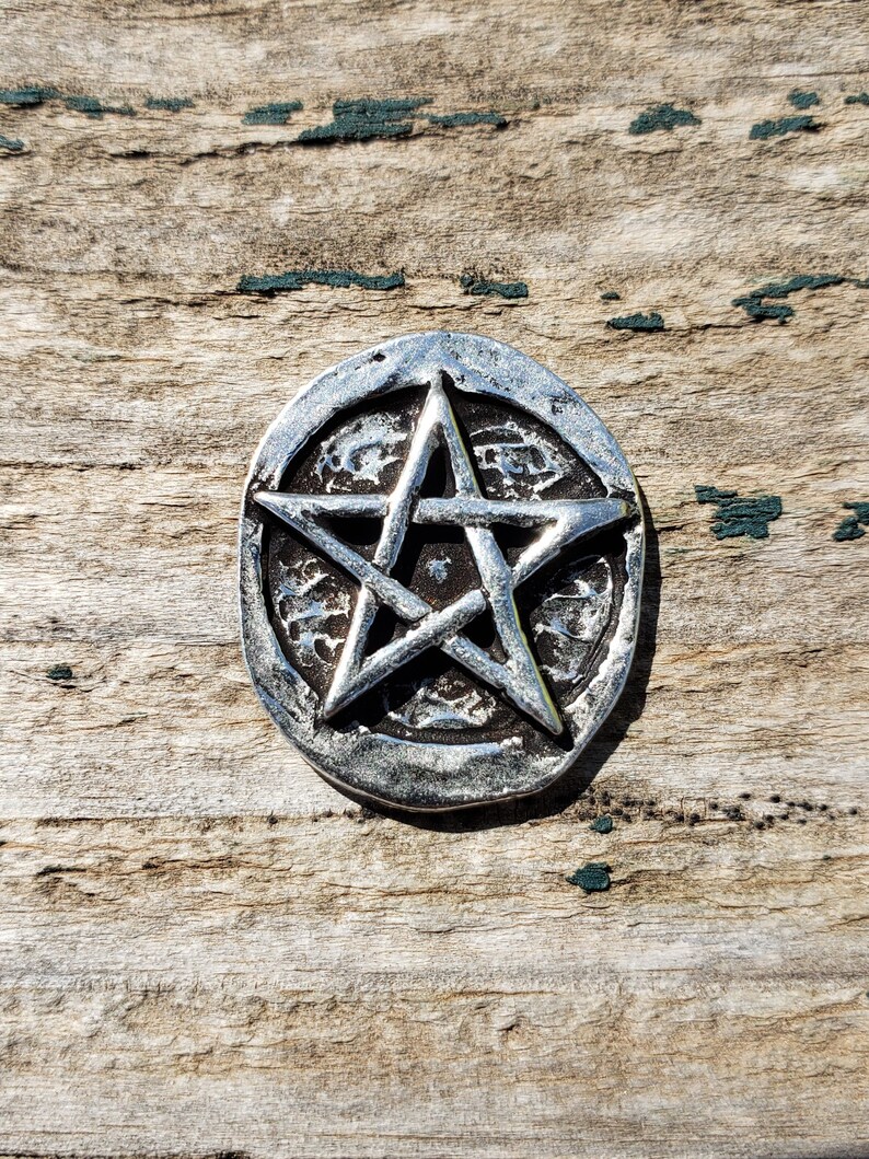 Puede incluir: Un colgante circular plateado con un dise&ntilde;o de pentagrama en relieve. El pentagrama es una estrella de cinco puntas dentro de un c&iacute;rculo, con un aspecto texturizado y envejecido. El colgante est&aacute; sobre una superficie de madera envejecida.