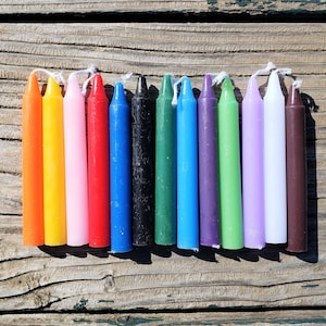 May include: A set of twelve cylindrical taper candles in a variety of solid colours including orange, yellow, pink, red, blue, black, green, light blue, purple, lime green, lavender, white, and brown, each featuring a white cotton wick at the top.
