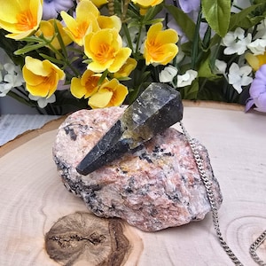 May include: A dark, faceted, pointed crystal pendant with a metallic chain resting on a rough, textured, pink and white mineral stone. The crystal features deep charcoal tones with subtle yellow and translucent inclusions throughout its structure.