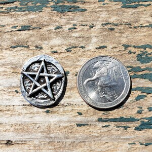 Puede incluir: Un colgante de pentagrama plateado con un dise&ntilde;o de estrella en relieve, junto a una moneda de veinticinco centavos estadounidense. El colgante es redondo con una superficie texturizada. El fondo es de madera envejecida con pintura turquesa descascarada.