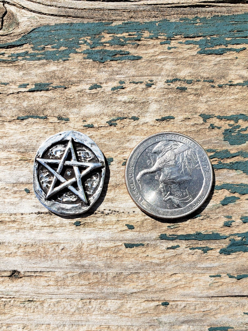 Puede incluir: Un colgante de pentagrama plateado con un dise&ntilde;o de estrella en relieve, junto a una moneda de veinticinco centavos estadounidense. El colgante es redondo con una superficie texturizada. El fondo es de madera envejecida con pintura turquesa descascarada.