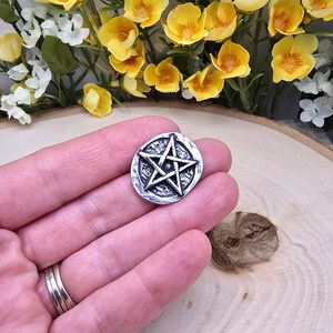 Puede incluir: Un colgante redondo plateado con un dise&ntilde;o de pentagrama en relieve. El colgante tiene un borde texturizado y se sostiene en una mano. El fondo incluye flores amarillas y blancas.