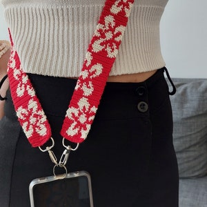 Puede incluir: Una correa de teléfono de crochet roja y blanca con un patrón floral. La correa está unida a una funda de teléfono transparente con una pantalla negra.