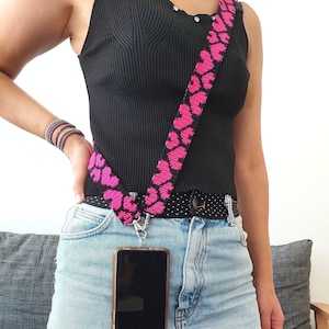 May include: A black tank top with a pink and black heart-shaped phone strap. The strap is attached to a black phone case with a clear back. The phone is in a denim skirt.