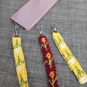 Pode incluir: Três tiras para telefone com designs florais. As tiras são amarelas, vermelhas e brancas com flores amarelas. As tiras são presas a chaveiros prateados.