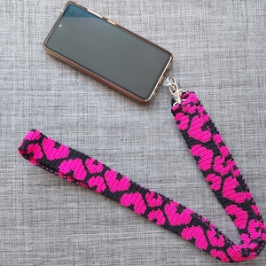 May include: A black phone case with a pink and black patterned phone strap. The strap has a metal clasp.