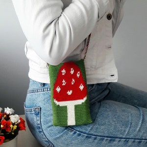 May include: A knitted mushroom-shaped phone case with a red top, white spots, and a white stem. The case is green on the bottom and is being worn by a person.