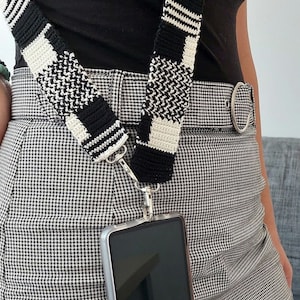 May include: A black and white crocheted phone strap with a silver clasp attached to a clear phone case. The phone case is attached to a black and white checkered skirt.