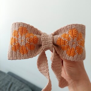 May include: A crocheted bow with a light pink base and orange floral accents. The bow has two long tails.