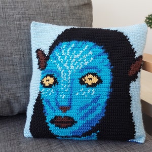 May include: A blue and black crocheted pillow with a close-up of a blue character's face. The character has large, yellow eyes and a brown nose.