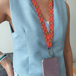 Puede incluir: Una correa de teléfono tejida de color rosa, verde y naranja con una funda de teléfono morada adjunta. La correa la lleva una persona que lleva una blusa sin mangas de color azul claro y pantalones vaqueros azules.
