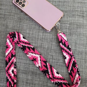 May include: A lavender smartphone with a gold-colored frame and a pink, black, and white patterned phone strap. The strap is made of woven material and is attached to the phone with a silver-colored clasp.