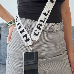 May include: A white and black crocheted phone strap with the words "CALL AGAIN" on it. The strap is attached to a clear phone case with a black smartphone inside. The phone is hanging from the strap and is worn by a person wearing a black top and a black and white checkered skirt.