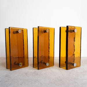 May include: Three rectangular amber glass wall sconces, each featuring two vertical glass panels held together by two circular silver-toned metal hardware pieces. The glass has a smooth, translucent finish with textured, slightly uneven edges.