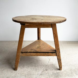 May include: A round wooden table with a weathered light brown finish, featuring three straight legs that taper slightly towards the floor and a triangular lower shelf positioned between the legs, showcasing a visible wood grain and textured surface.