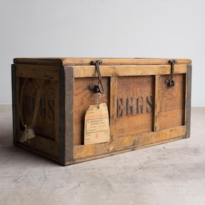 May include: A rectangular wooden crate with dark metal corner reinforcements and two metal latches on the front. The word EGGS is printed in black on the front and side. A beige shipping tag labelled To BARNSLEY is attached to a latch.