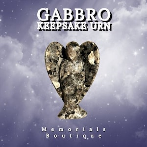 May include: A gabbro keepsake urn in the shape of an angel with outstretched wings. The angel is carved from a speckled gray and tan stone. The text "Gabbro Keepsake Urn" is at the top, and "Memorials Boutique" is at the bottom.