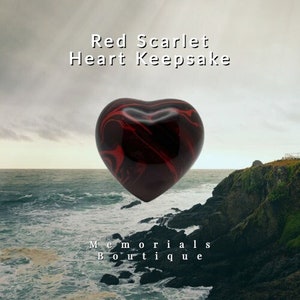 May include: Red heart-shaped keepsake made of red and black marbled resin. The heart is displayed against a backdrop of a cloudy sky and a rocky coastline with ocean waves.