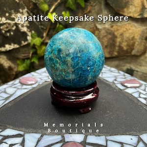 May include: Apatite Keepsake Sphere. A blue-green sphere sits on a dark red wooden stand. The sphere has a textured surface with a mix of blue and green tones. The text "Memorials Boutique" is at the bottom.