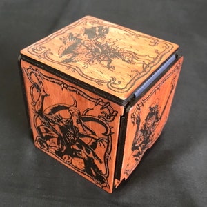 May include: A wooden box with a black and brown finish. The box is decorated with an intricate design of a creature with wings and claws. The box is made of six panels that fit together to form a cube.