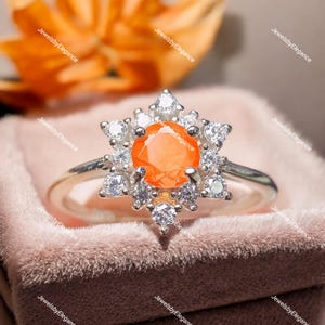 May include: A silver ring featuring a vibrant orange gemstone at the center, surrounded by a halo of smaller, clear gemstones. The ring's design is floral-inspired, with the gemstones arranged to resemble petals. The ring is displayed on a pink velvet surface.