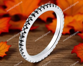 May include: A silver ring featuring a continuous row of small, black gemstones. The ring is set against a blurred backdrop of autumnal leaves and a wooden surface. The ring is marked with the text "JewelsbyElegance".