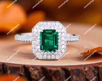 May include: A close-up of a silver ring with a large emerald-cut, deep green gemstone, surrounded by a halo of smaller, clear stones. The ring is set on a wooden surface, with the text "JewelsbyElegance" repeated around the image.