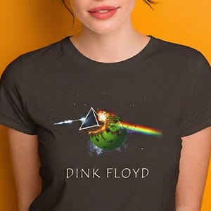May include: Dark gray t-shirt featuring a graphic of a prism refracting light into a rainbow, with a planet in the center. The text "DINK FLOYD" is printed below the graphic. The shirt is worn by a person with brown hair and blue eyes.