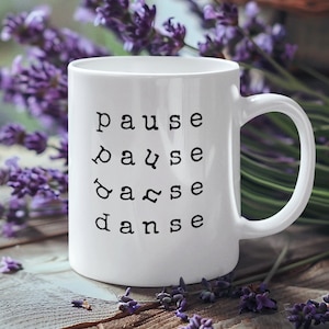 Könnte beinhalten: Eine weiße Keramiktasse mit einer schwarzen Aufschrift, die "pause,  pause,  dance,  dance" lautet. Die Tasse steht auf einer Holzoberfläche mit violetten Lavendelblüten.