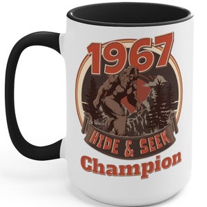May include: White ceramic mug with a black handle and black rim. The mug features a brown Bigfoot illustration with the text "1967 Hide & Seek Champion" in a vintage style.