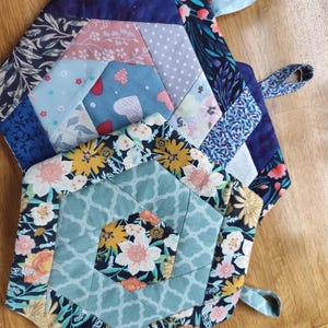 May include: Three hexagonal quilted pot holders. They display a variety of floral and geometric patterns in shades of blue, pink, and yellow. Each pot holder has a small loop for hanging, perfect for a kitchen.