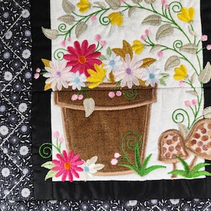 May include: Embroidered floral design featuring a brown flower pot overflowing with colourful flowers. The design includes pink, yellow, and white flowers, green leaves, and two mushroom accents. The border is black and patterned.