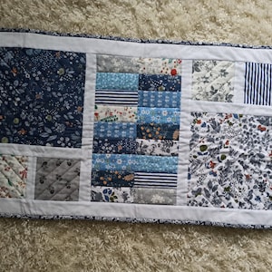 May include: A long, rectangular table runner with a patchwork design. The runner is made of various blue and white fabrics with floral and geometric patterns. The runner is edged with a white border.