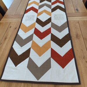 May include: A rectangular table runner featuring a repeating chevron pattern in shades of burnt orange, mustard yellow, brown, beige, and cream. The design includes two square panels with intricate floral embroidery and a dark brown border trim.
